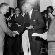 grupa odświętnie ubranych osób w średnim wieku stojąca w zabytkowym pomieszczeniu na tle okna z kratownicą; na ścianach widoczne zdobienia i świece