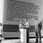 mężczyzna w garniturze stojący przy pulpicie z flagą Polski i godłem PRL, przemawiający do mikrofonu, obok stoją dwaj inni mężczyźni, jeden z nich trzyma drugi mikrofon 