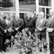 grupa mężczyzn w garniturach i jedna kobieta w garsonce na tle jednego z pawilonów targowych