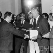 grupa odświętnie ubranych osób w średnim wieku stojąca w zabytkowym pomieszczeniu na tle okna z kratownicą; na ścianach widoczne zdobienia i świece