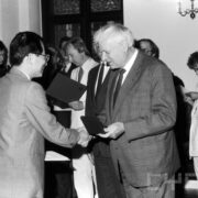 grupa odświętnie ubranych osób w średnim wieku stojąca w zabytkowym pomieszczeniu na tle okna z kratownicą; na ścianach widoczne zdobienia i świece