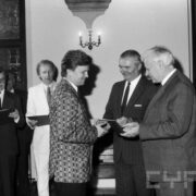 grupa odświętnie ubranych osób w średnim wieku stojąca w zabytkowym pomieszczeniu na tle okna z kratownicą; na ścianach widoczne zdobienia i świece