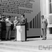 mężczyzna w garniturze stojący przy pulpicie z flagą Polski i godłem PRL, przemawiający do mikrofonu, wokół stoją inni mężczyźni, jeden z nich trzyma kamerę, kilku mikrofony 