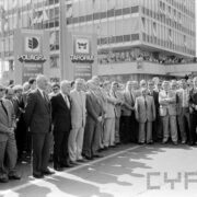 mężczyźni w garniturach stojący na placu na tle przeszklonego wieżowca