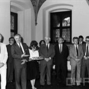 grupa odświętnie ubranych osób w średnim wieku stojąca w zabytkowym pomieszczeniu na tle okna z kratownicą; na ścianach widoczne zdobienia i świece