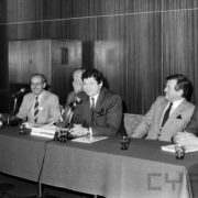 pięciu mężczyzn w średnim wieku, ubranych w garnitury siedzi za stołem, na którym stoją mikrofony, szklanki i dekoracje