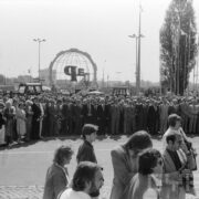 tłum ludzi stojących na dużym placu, w tle widoczne maszty i banery reklamowe
