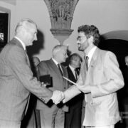 grupa odświętnie ubranych osób w średnim wieku stojąca w zabytkowym pomieszczeniu na tle okna z kratownicą; na ścianach widoczne zdobienia i świece