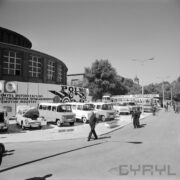 stoisko z samochodami na poboczu ulicy, z lewej murowany okrągły budynek z oknami, przed samochodami spacerujący ludzie