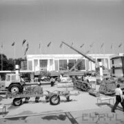 maszyny rolnicze i samochody na placu, w głębi murowany budynek