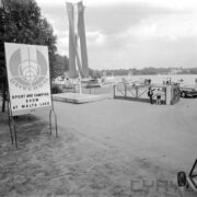 wjazd przez bramę na teren wokół jeziora, z lewej rozłożone namioty, spacerujący ludzie, z lewej stojąca reklama