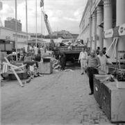 rozładunek skrzyń z samochodu ciężarowego, z prawej grupa mężczyzn i budynek z kolumnami