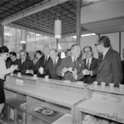 grupa mężczyzn stoi przy ladzie na której ustawiono filiżanki, część z mężczyzn trzyma w rękach filiżanki, z lewej kobieta ze szklanym dzbankiem