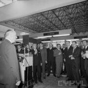 grupa mężczyzn w pawilonie wystawowym, z lewej mężczyzna przy mikrofonie