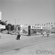 zadaszone stoisko na betonowym placu, w głębi murowany budynek, spacerujący ludzie, na pierwszym planie tory kolejowe