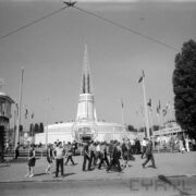 maszty z flagami państw i ażurową konstrukcją kuli ziemskiej pośrodku, w głębi okrągły budynek z ażurową iglicą na dachu, spacerujący ludzie