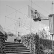 kobieta w koszu na podnośniku maluje napis na metalowej konstrukcji, poniżej schody kamienne i murek obrośnięty pnączem