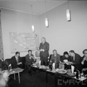 grupa mężczyzn i kobiet siedząca przy stołach, na stołach szklanki, kieliszki, butelki i talerze