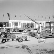 maszyny rolnicze i samochody na placu, w głębi murowany budynek