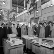 grupa ludzi w pawilonie wystawowym, z przodu stoisko z elektrycznymi maszynami liczącymi