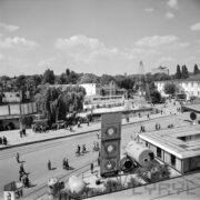 ulica z torami kolejowymi na terenie wystawy, dookoła budynki wystawowe, drzewa, spacerujący ludzie