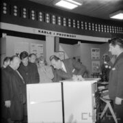 grupa ludzi, jeden z mężczyzn prezentuje im wnętrze otwartej lodówki 