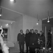 grupa mężczyzn i jedna kobieta, w większości w płaszczach, w hali wystawowej