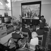 trzech mężczyzn siedzi przodem do widowni siedzącej na krzesłach w pawilonie wystawowym, na ścianie duże zdjęcie przywódcy koreańskiego w otoczeniu dzieci