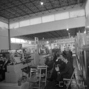 grupa mężczyzn siedzi na krzesłach w pawilonie wystawowym, obok stoją maszyny i urządzenia