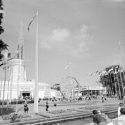 maszty z flagami państw i ażurową konstrukcją kuli ziemskiej pośrodku, w głębi okrągły budynek z ażurową iglicą na dachu, na placu spacerujący ludzie