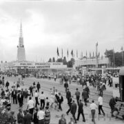 ludzie spacerujący ulicą na terenie wystawy, środkiem biegną tory kolejowe, w głębi budynki, z lewej okrągły budynek z ażurową iglicą na dachu