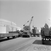 ulica na terenie targów, na niej samochody dostawcze z przyczepami oraz dźwig samochodowy, z lewej fragment okrągłego budynku