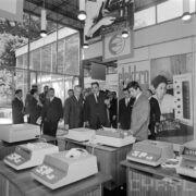 grupa ludzi w pawilonie wystawowym, z przodu stoisko z elektrycznymi maszynami liczącymi