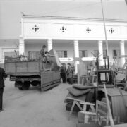 dwóch mężczyzn ściąga z przyczepy samochodu dostawczego skrzynię, dwóch mężczyzn stoi obok, w głębi otynkowany budynek z kolumnami, z prawej poukładane na stosie meble