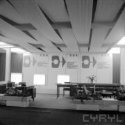 z lewej biurko drewniane, z prawej stolik z fotelami, za nimi dwa biurka, w głębi tablice reklamowe