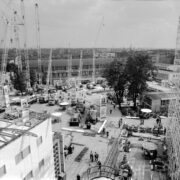 plac z dźwigami i maszynami budowlanymi, dookoła budynki wystawowe, spacerujący ludzie