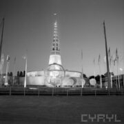 murowany budynek z iglicą, przed nim ażurowy model kuli ziemskiej z literką P, po obu stronach maszty z flagami państw, całość sfotografowana nocą