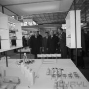 kilku mężczyzn, część z nich w płaszczach, oglądają stoisko w hali targowej