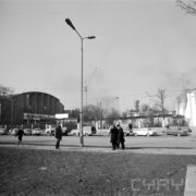 ulica ze stojącymi wzdłuż niej zaparkowanymi samochodami, na pierwszym planie idzie chodnikiem troje ludzi w płaszczach: trzech mężczyzn i jedna kobieta, w głębi tereny targowe, z lewej fragment murowanej hali targowej, pośrodku lampa uliczna