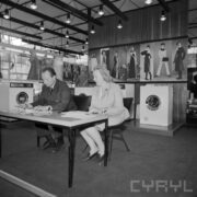 kobieta i mężczyzna siedzą przy stoliku, za nimi stoisko z pralkami