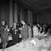 grupa kobiet i mężczyzn stoją między dwoma rzędami nakrytych obrusem i zastawionych stołów