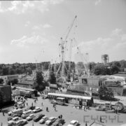 na pierwszym planie aleja na terenie targów, spacerujący ludzie, w głębi budynki wystawowe, i dźwigi budowlane, na placu zaparkowane samochody