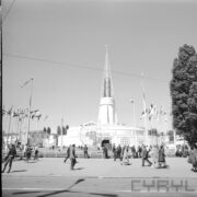 maszty z flagami państw i ażurową konstrukcją kuli ziemskiej pośrodku, w głębi okrągły budynek z ażurową iglicą na dachu, na placu spacerujący ludzie