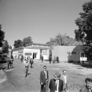 spacerujący ludzie po terenach targowych, w głębi murowany budynek 