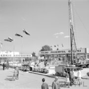 plac z wystawionymi maszynami, na pierwszym planie spacerują ludzie, w głębi dwa budynki, na placu maszty z powiewającymi falagami