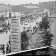 ulica z torami kolejowymi na terenie wystawy, dookoła budynki wystawowe, drzewa, spacerujący ludzie