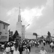 tłumy ludzi spacerujący po alei na terenie targów, w głębi okrągły budynek z ażurową iglicą na dachu