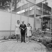 mężczyzna w środku, pod rękę z obu stron trzyma dwie kobiety, cała trójka pozuje na tle przeszklonego budynku