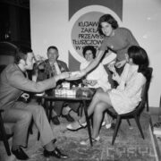 z prawej dwie kobiety siedzą na fotelach, jedna stoi pomiędzy nimi, z lewej dwaj mężczyźni siedzą na fotelach, na środku stoi mały stolik