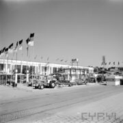 stoisko z maszynami rolniczymi na placu, w głębi budynek murowany, z lewej maszty z powiewającymi flagami Niemieckiej Republiki Demokratycznej